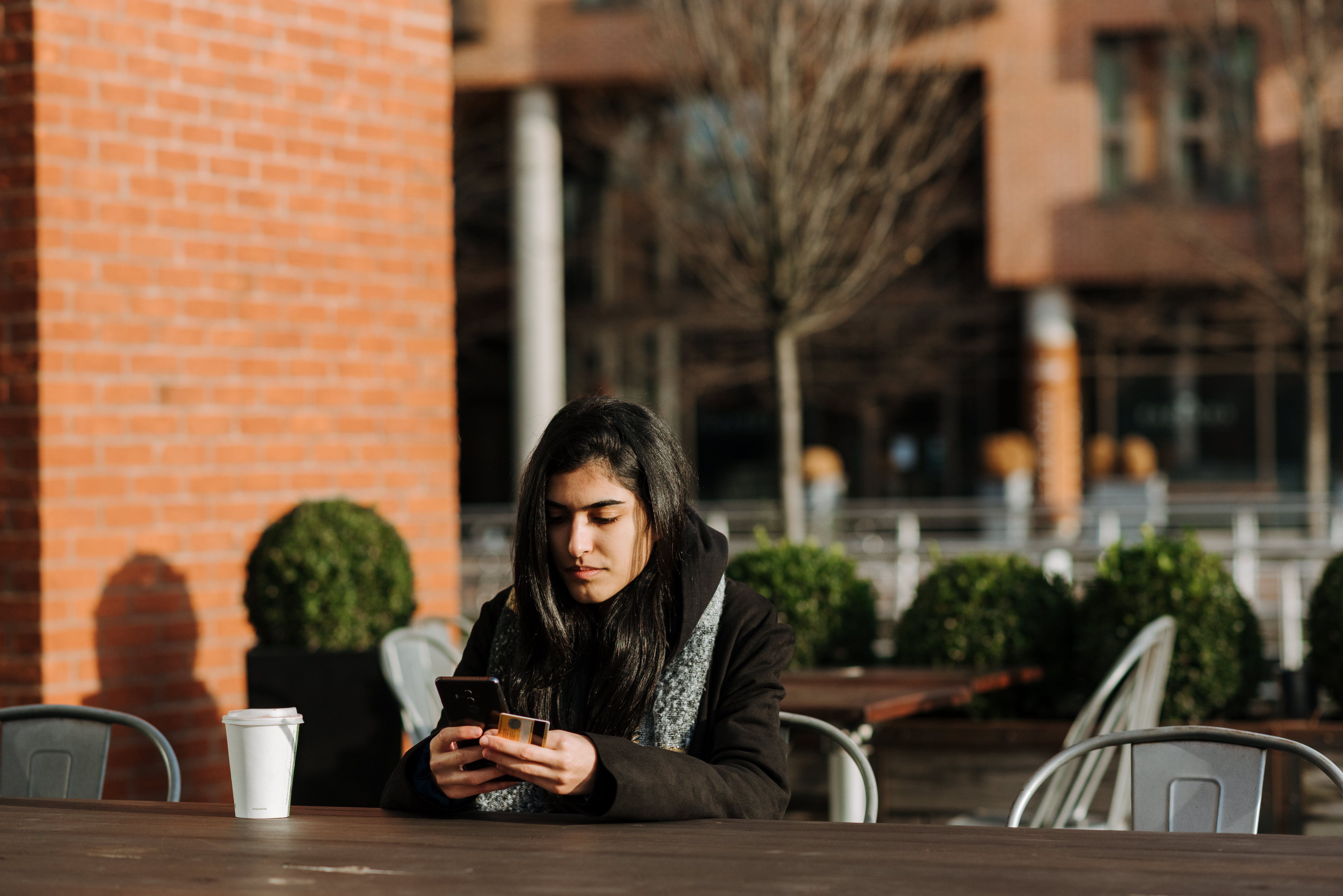 A person sitting alone at a café table, looking at their phone, coat still on