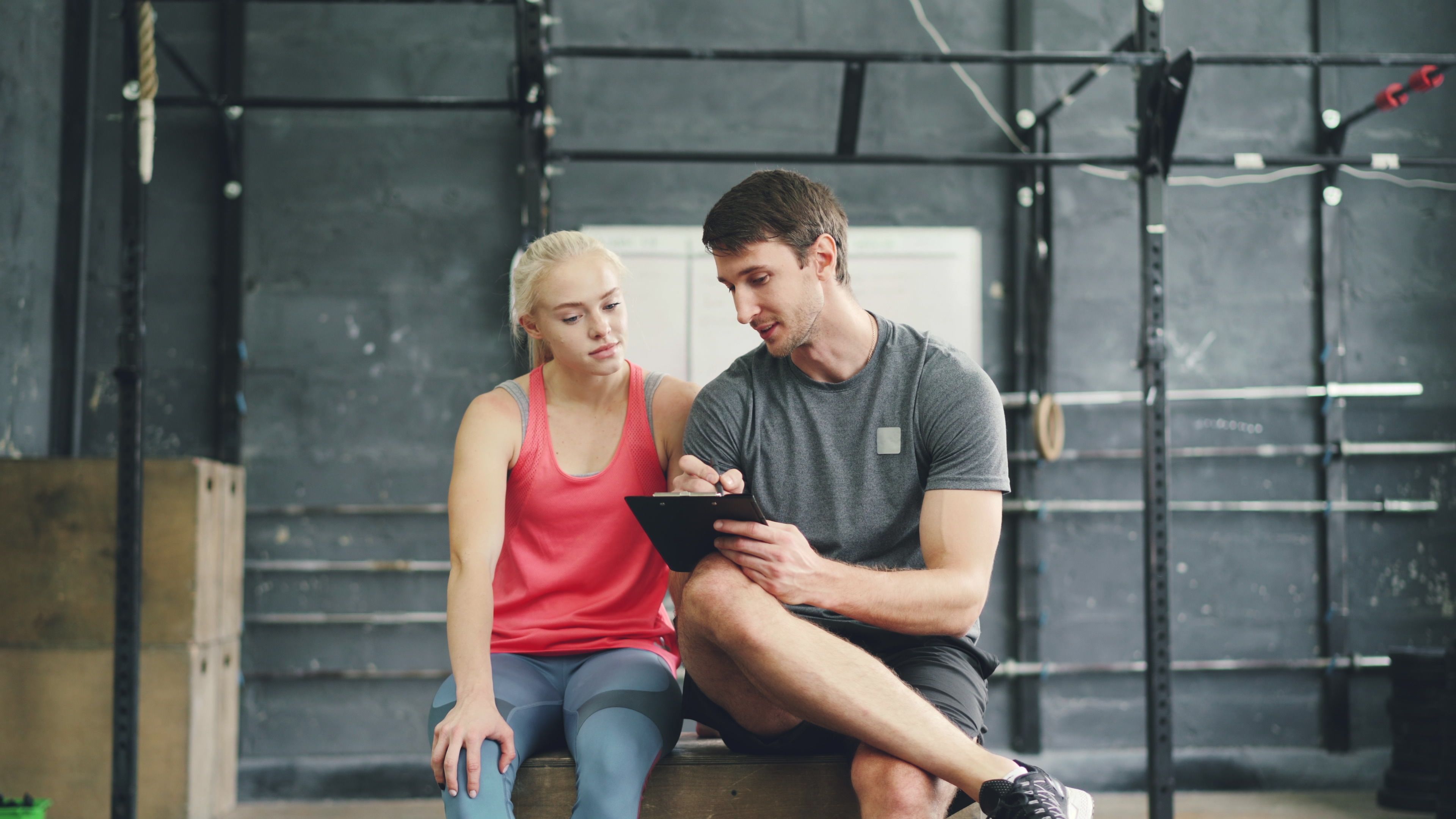 A coach and a client talking at the side of a gym floor, clipboard down, mid-conversation